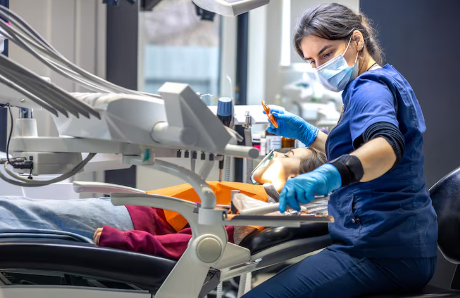 Female dentist wearing a mask and gloves treating a young patient in a modern dental clinic while using dental tools and equipment.